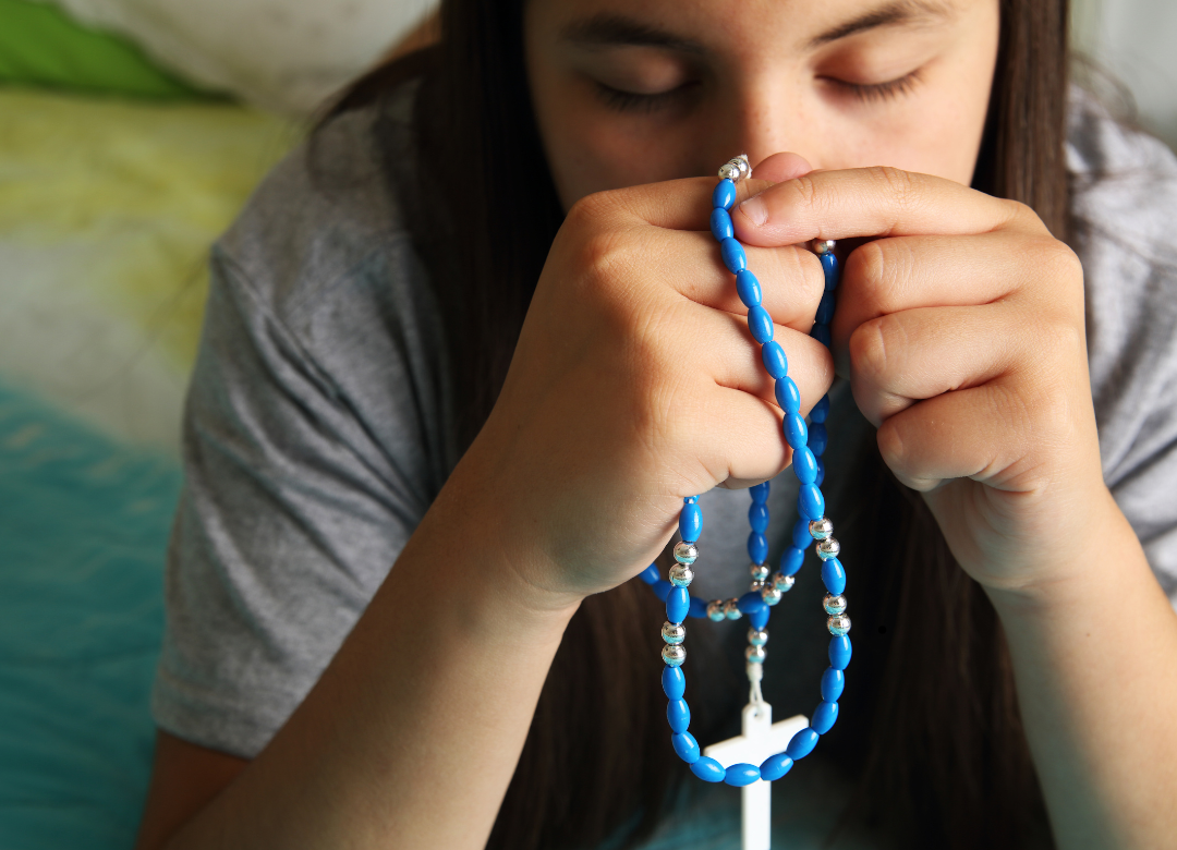 A teenage girl prays with her eyes closed, holding a blue rosary. The rosary is a spiritual tool Catholic students can use to grow closer to the Virgin Mary. (via Canva)