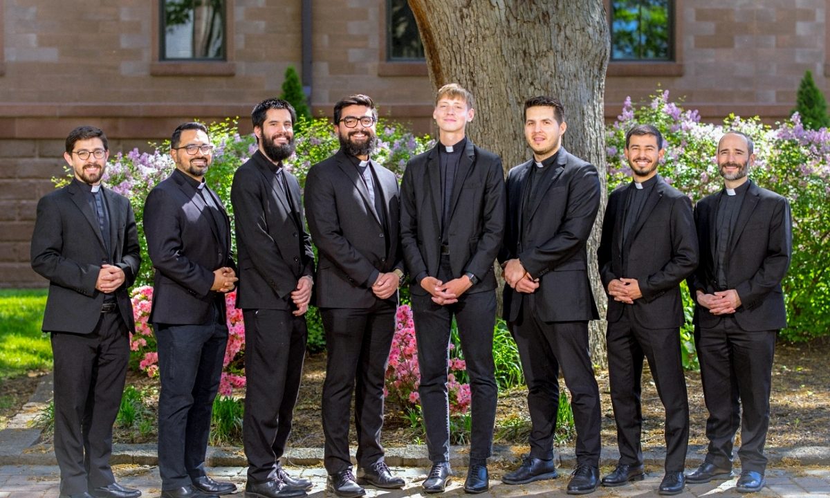A group of eight men stand together in front of a tree outside.