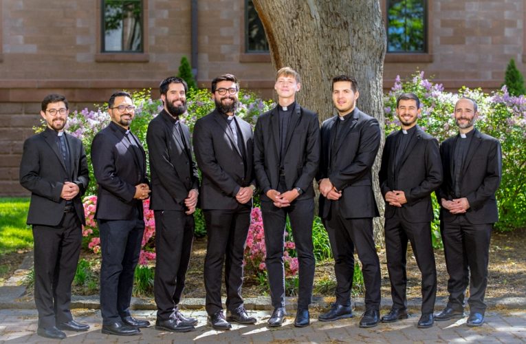 A group of eight men stand together in front of a tree outside.